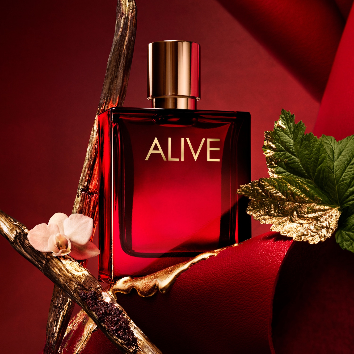 Czerwony flakon perfum Alive z złotym napisem na tle dekoracji z orchidei, liści i gałązek.