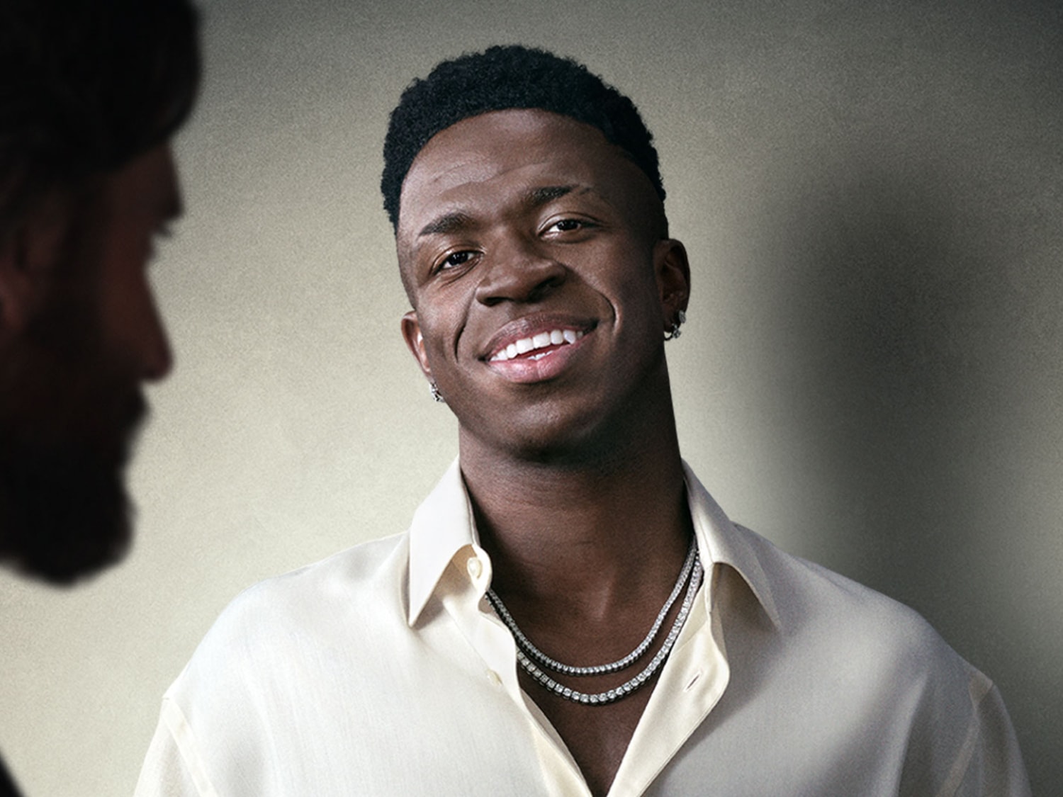 Portret uśmiechniętego mężczyzny o ciemnej karnacji w koszuli w kolorze złamanej bieli i dwóch diamentowych łańcuszkach na szyi, z zarysem brodatej twarzy po lewej stronie.