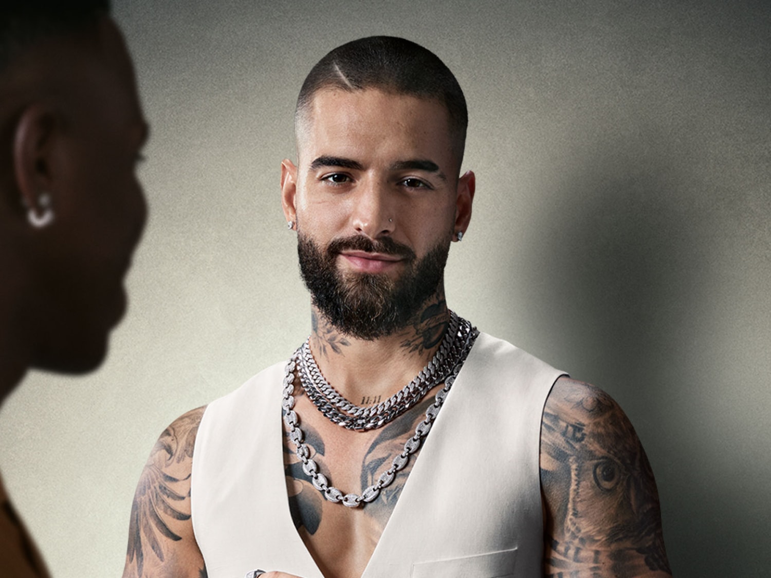 Portret Malumy, ubranego w białą kamizelkę i srebrne łańcuszki, z tatuażami na szyi i ramionach, spoglądającego w obiektyw z lekkim uśmiechem.