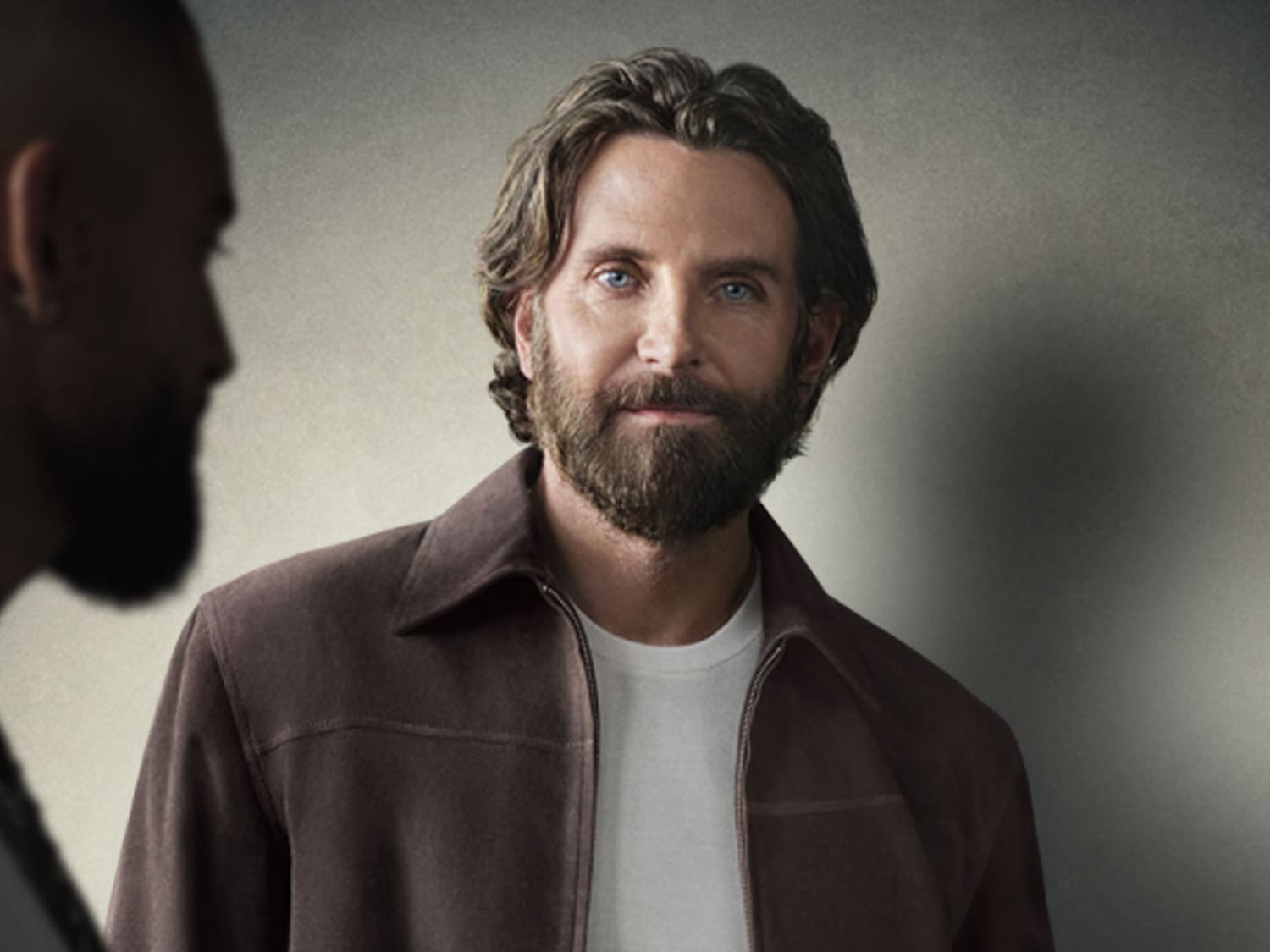 Portret mężczyzny z brodą i niebieskimi oczami, ubranego w brązową kurtkę i białą koszulkę, z częścią twarzy innego mężczyzny widoczną po lewej stronie.