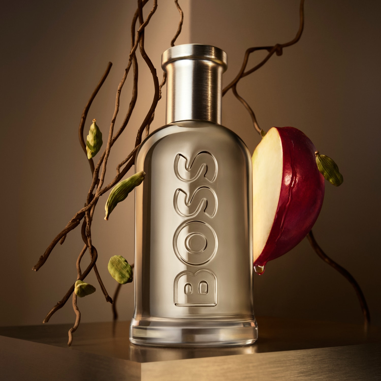 Błyszczący, srebrny flakon perfum BOSS w towarzystwie gałązek i soczystego kawałka czerwonego jabłka, tworząc elegancką kompozycję.