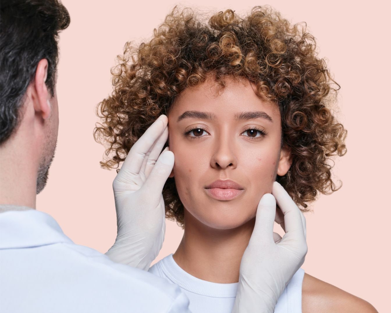 Un dermatologue examine la peau d'une jeune femme aux cheveux bouclés, portant des gants blancs pour un examen facial professionnel.