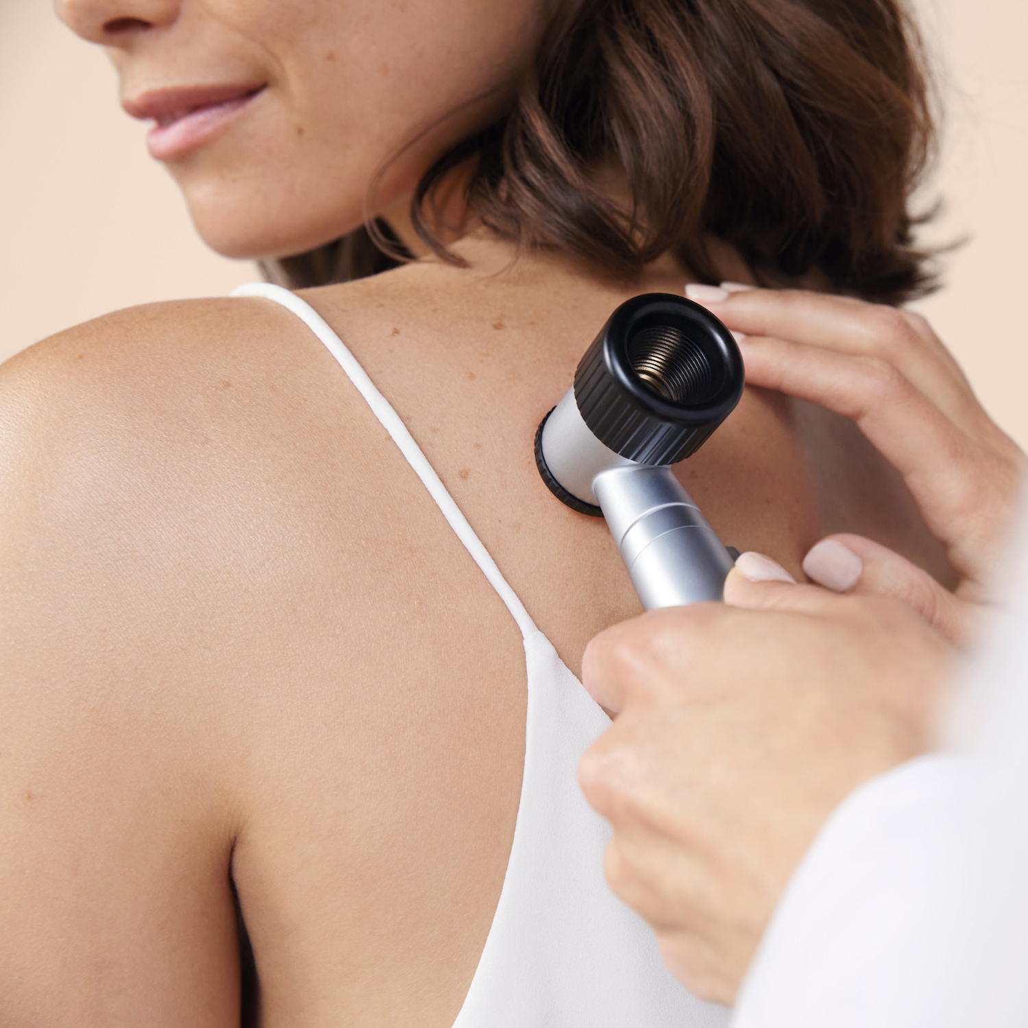 Une femme en débardeur blanc se fait examiner le dos avec un dermatoscope argenté et noir pour vérifier ses grains de beauté.