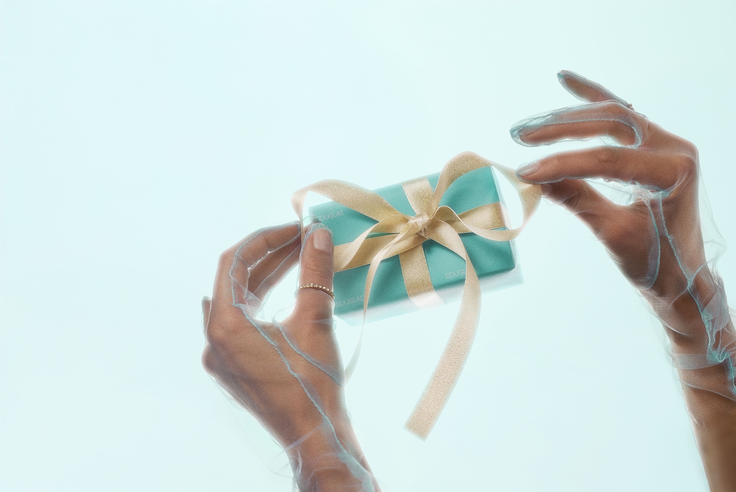 Twee handen met doorschijnende handschoenen maken een gouden lint los van een turquoise Douglas-geschenkdoos.