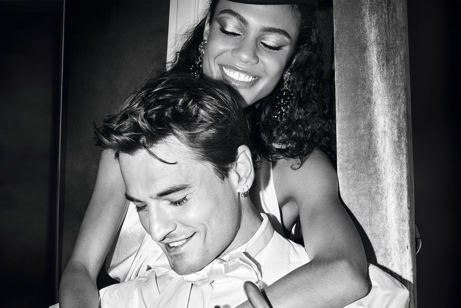 Een zwart-wit foto van een lachende vrouw met krullend haar en glinsterende oogschaduw die een man omhelst, beiden met hun ogen gesloten.