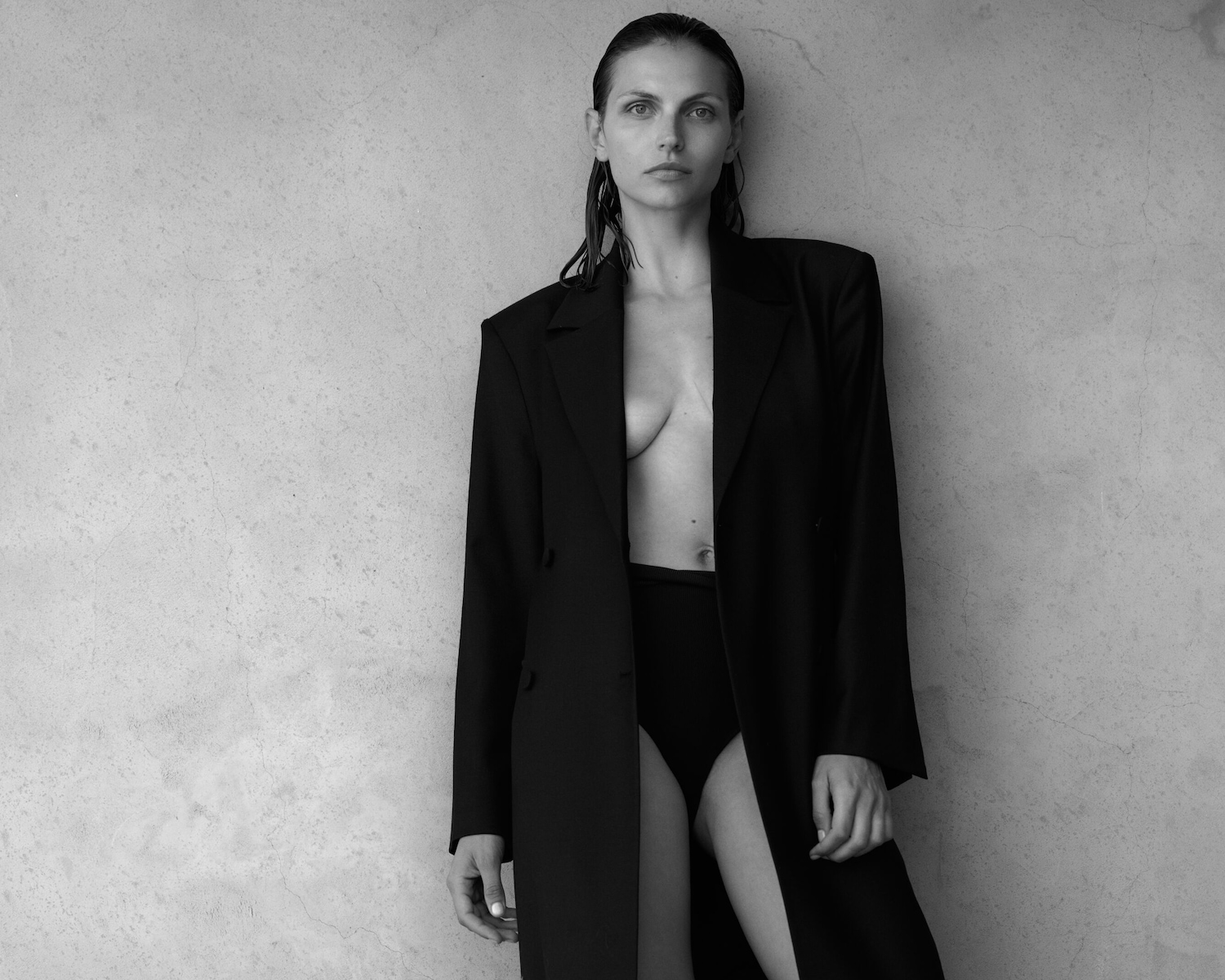 Schwarz-Weiß-Aufnahme einer Frau mit nassen Haaren, die einen offenen schwarzen Blazer und einen schwarzen Slip vor einer strukturierten Wand trägt.