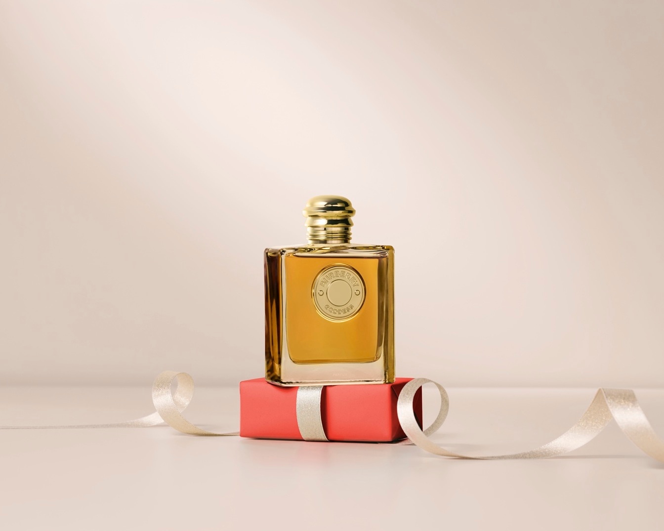 Un flacon de parfum Burberry Goddess doré repose sur un cadeau rouge corail orné d'un ruban argenté, le tout sur un fond beige doux.