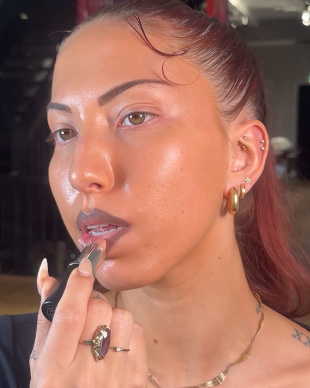 Een vrouw brengt lippenstift aan, met een zwarte verpakking, terwijl ze gouden sieraden draagt, waaronder oorbellen, een ketting en een ring.