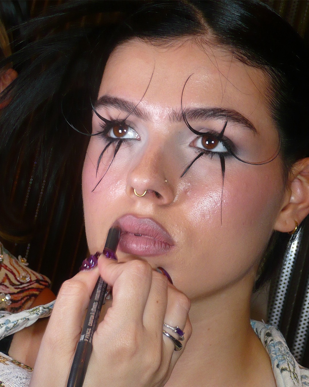 Een vrouw met een gouden neusring en opvallende wimpers krijgt lippenstift aangebracht met een donkerbruin potlood.