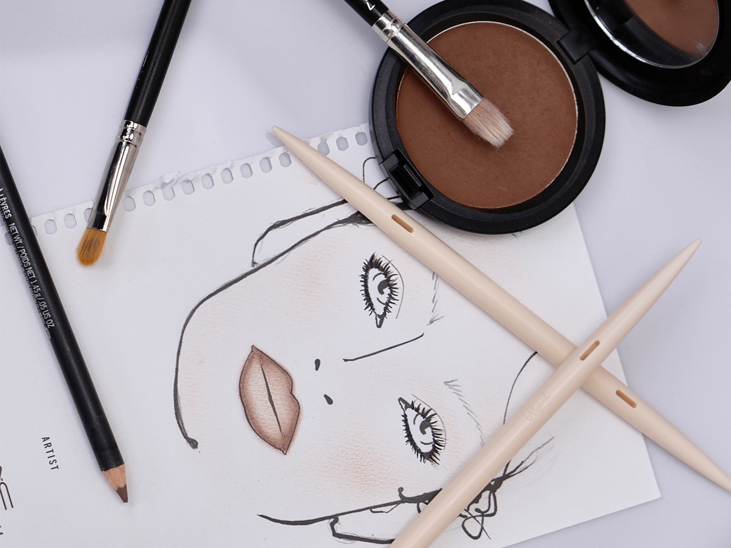 Een make-upschets met penselen, een bruin poeder en potloden, ideaal voor het plannen van je volgende beauty look.