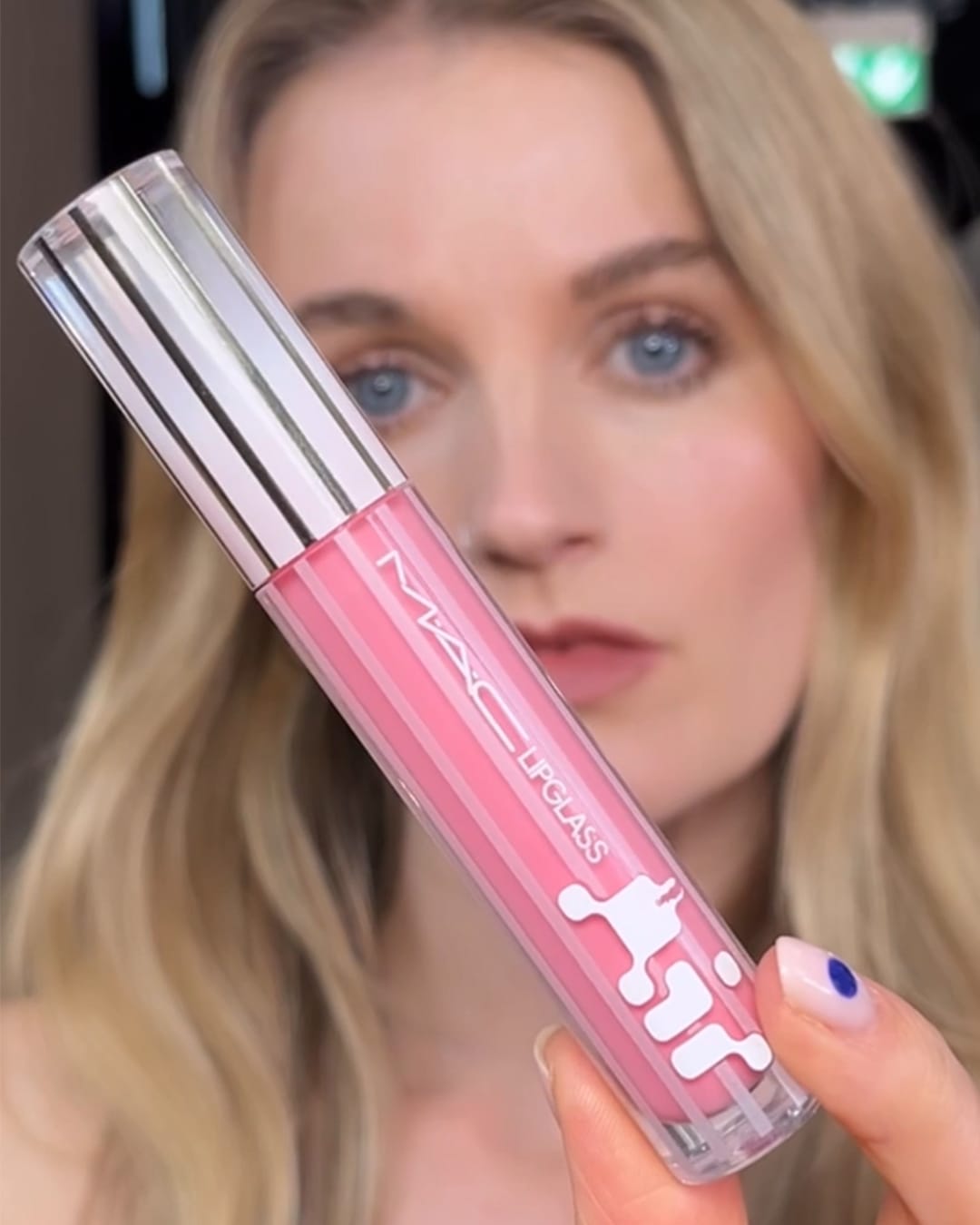Een blonde vrouw houdt een MAC Lipglass vast, de verpakking is roze met witte details en de dop is zilverkleurig.