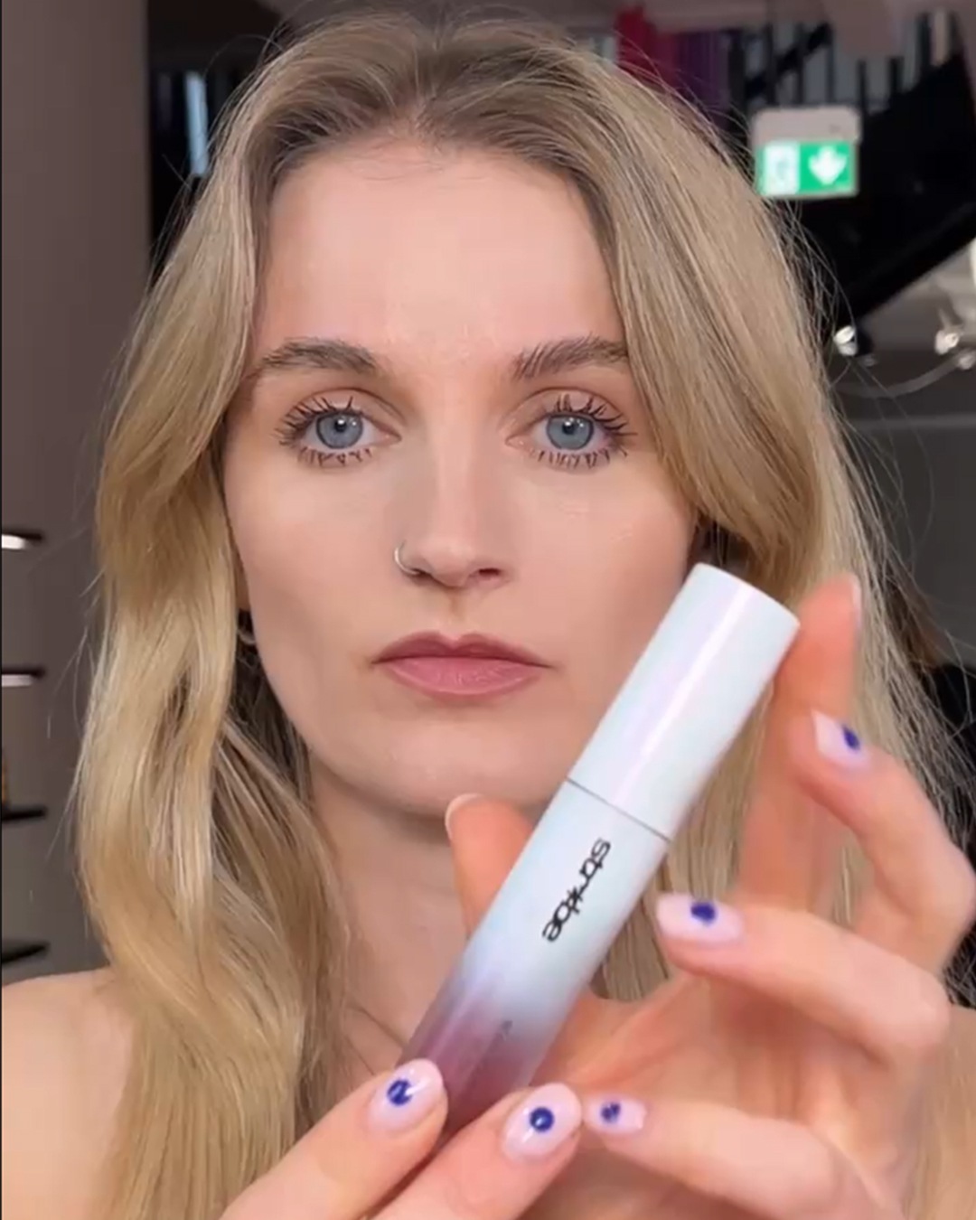 Een vrouw met blond haar houdt een Starloe mascara vast, de verpakking is wit en loopt over in een paarse kleur.