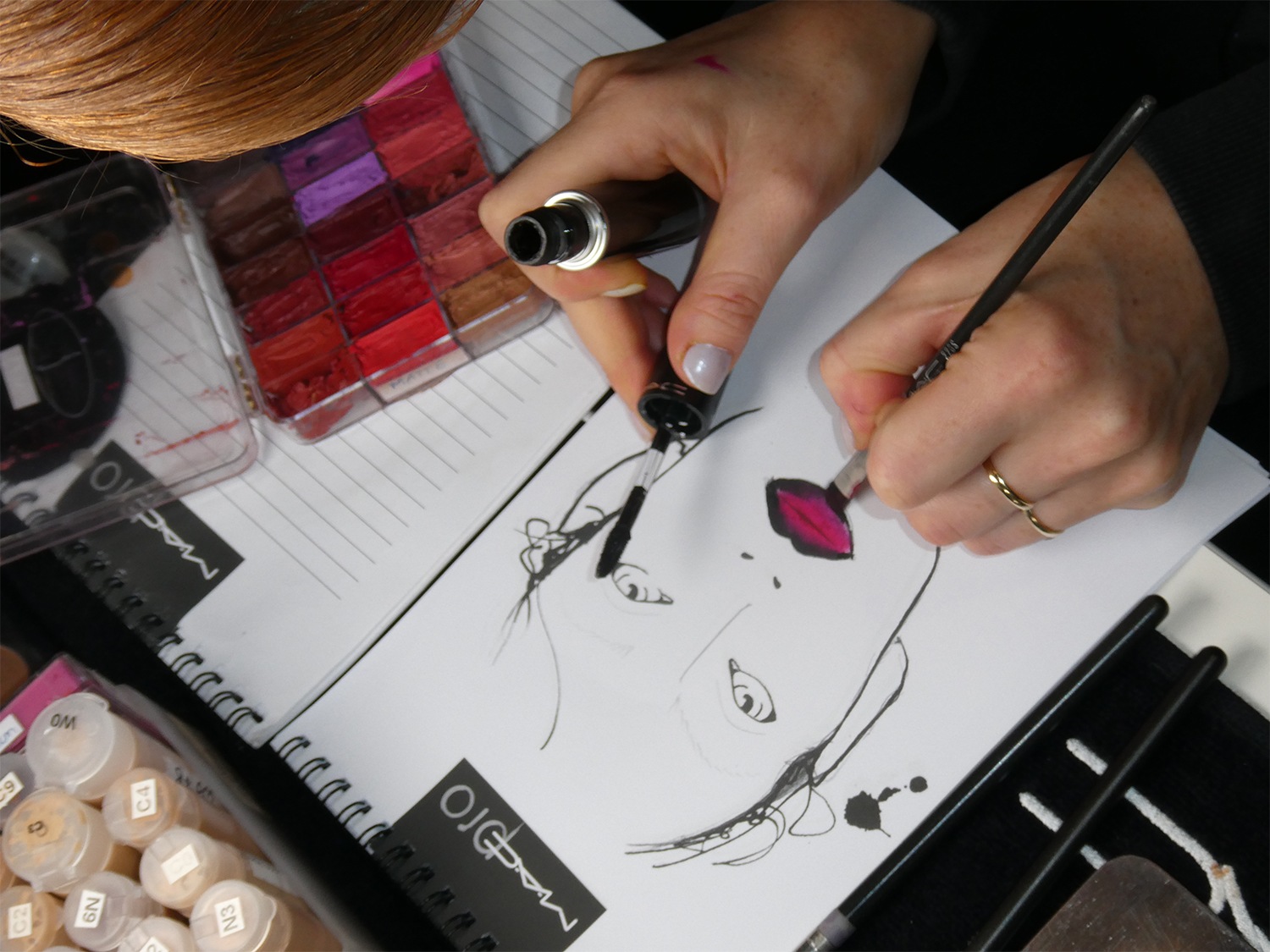 Een visagist tekent een gezicht op papier en gebruikt een penseel om de lippen roze te kleuren, met een palet aan kleuren en make-up potjes in de buurt.