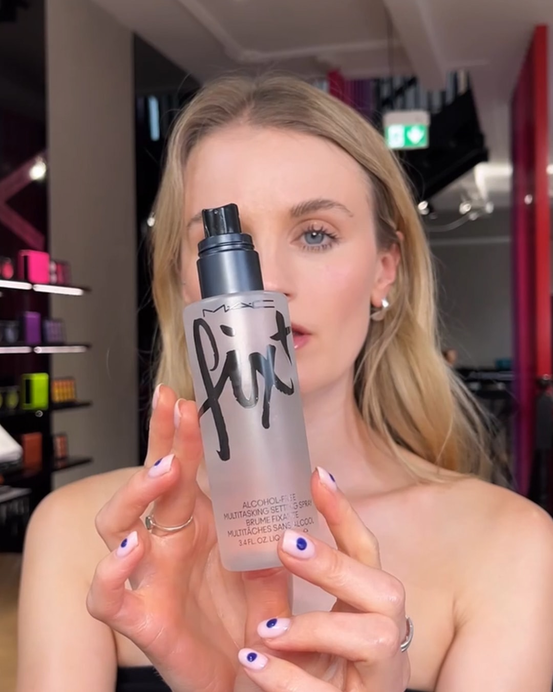 Een blonde vrouw houdt een transparante fles M.A.C. Fix+ vast, een alcoholvrije, multitaskende setting spray.
