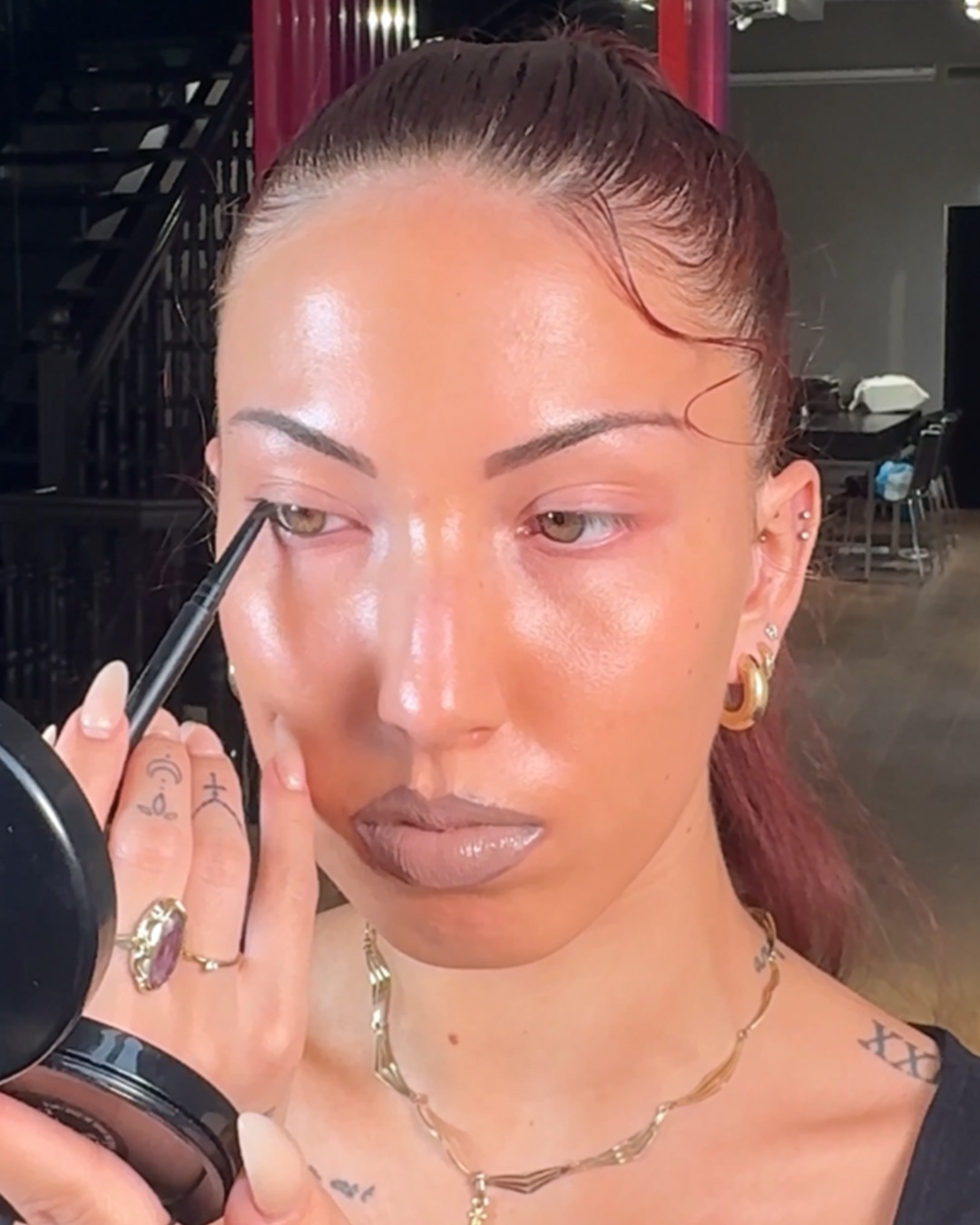 Een vrouw brengt eyeliner aan met een zwart potlood, kijkend in een compacte spiegel, met een gouden ketting en ringen als detail.