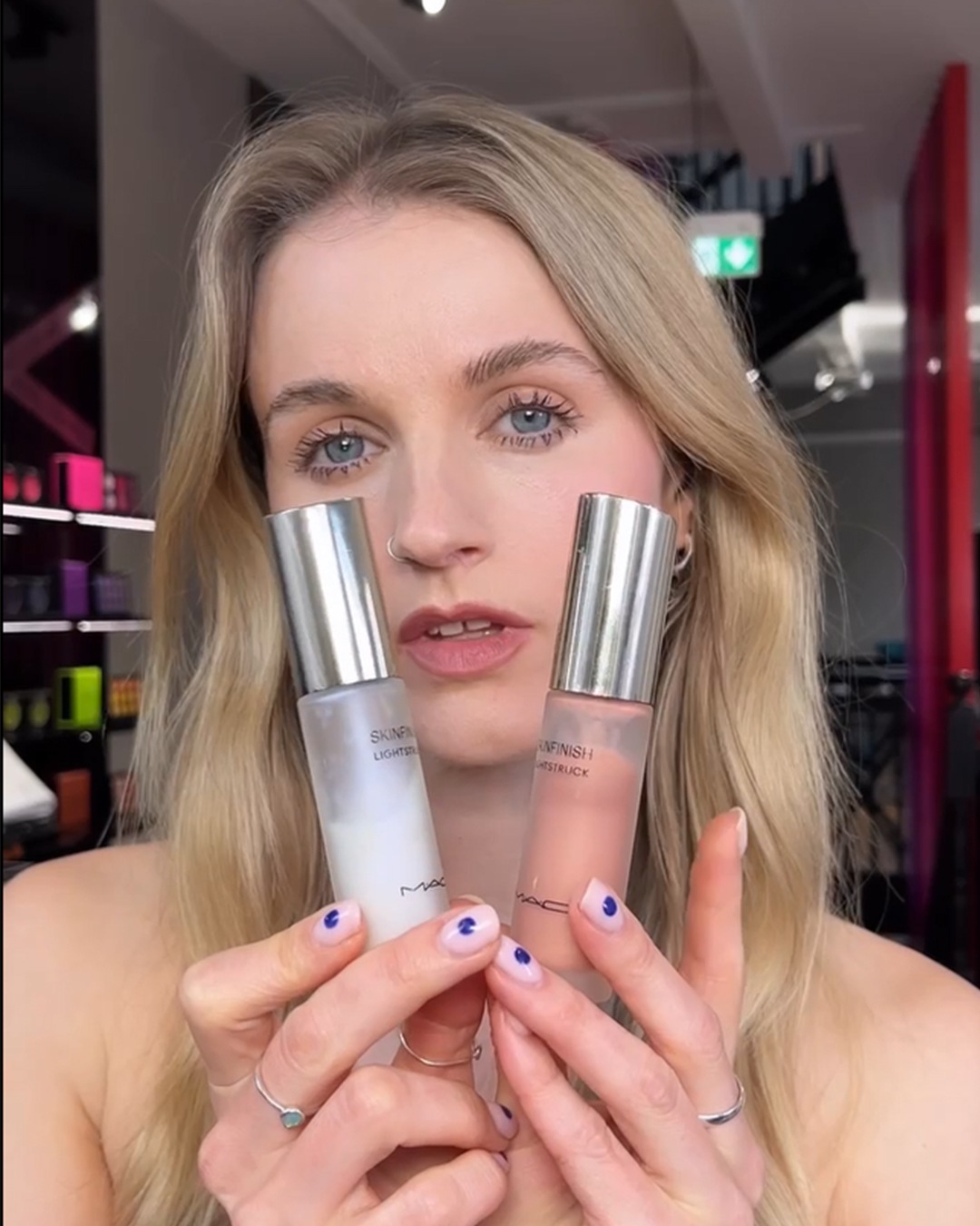 Een blonde vrouw toont twee MAC-sprays, de ene Skinfinish Lightstruck in een witte fles en de andere Skinfinish Heatstruck in een perzikkleurige fles.