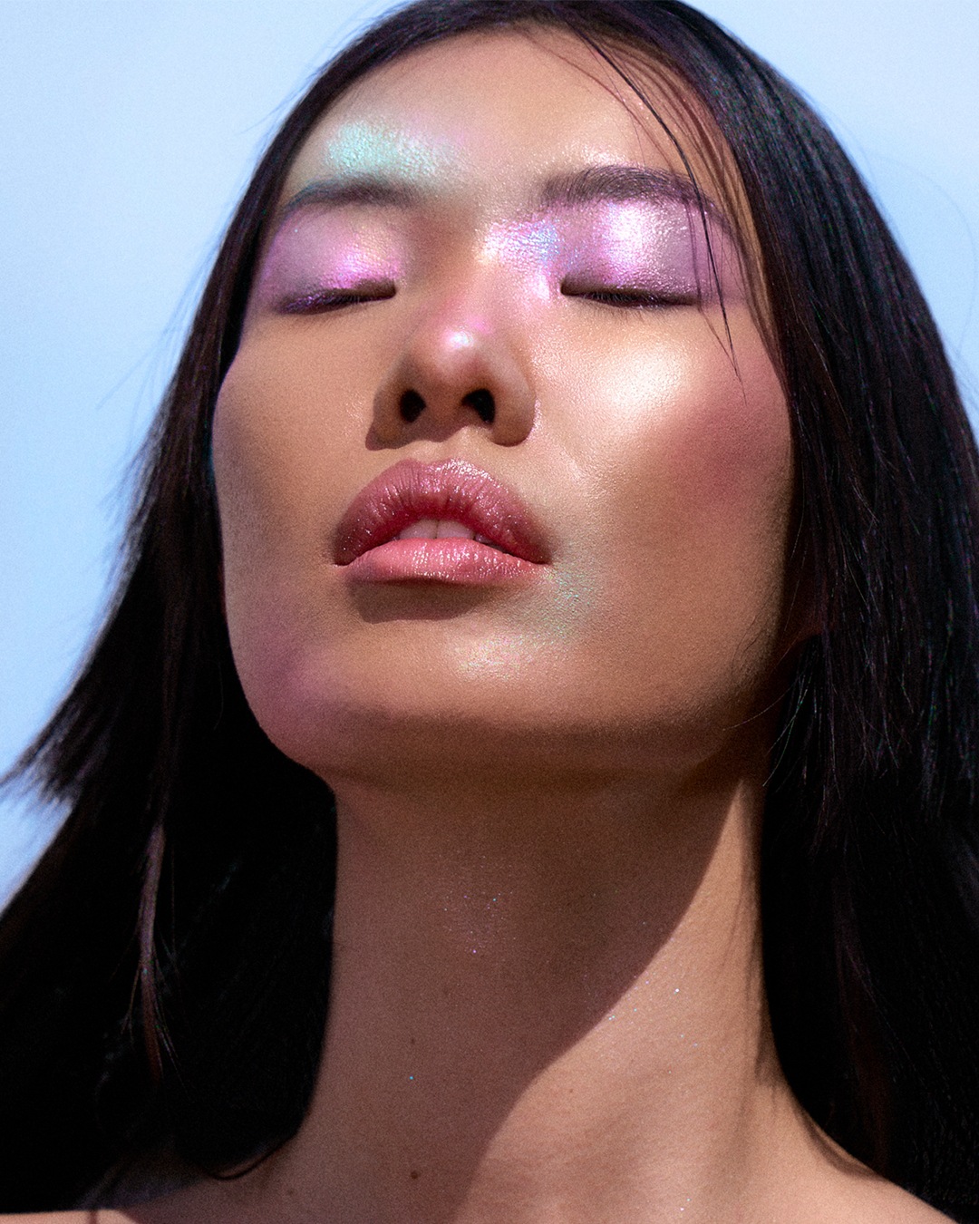 Een vrouw met gesloten ogen, glanzende roze en paarse oogschaduw, en glanzende lippenstift, haar huid heeft een iriserende glans.