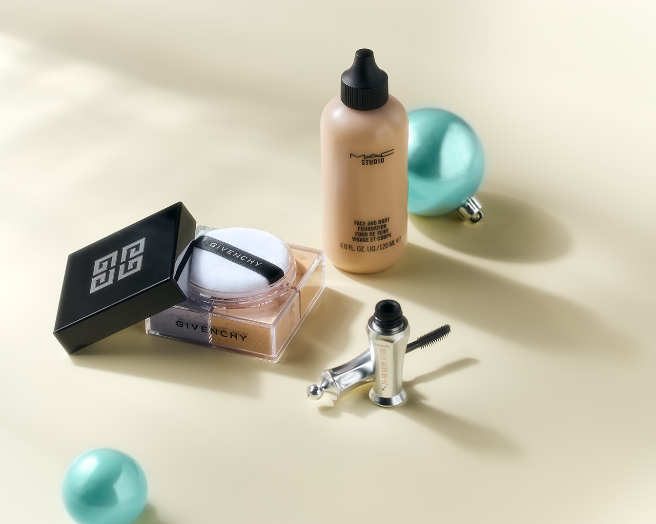 Découvrez un assortiment de produits de beauté, comprenant la poudre libre Givenchy, le fond de teint visage et corps Studio de MAC et le mascara sourcils Benefit, présentés avec une décoration festive.