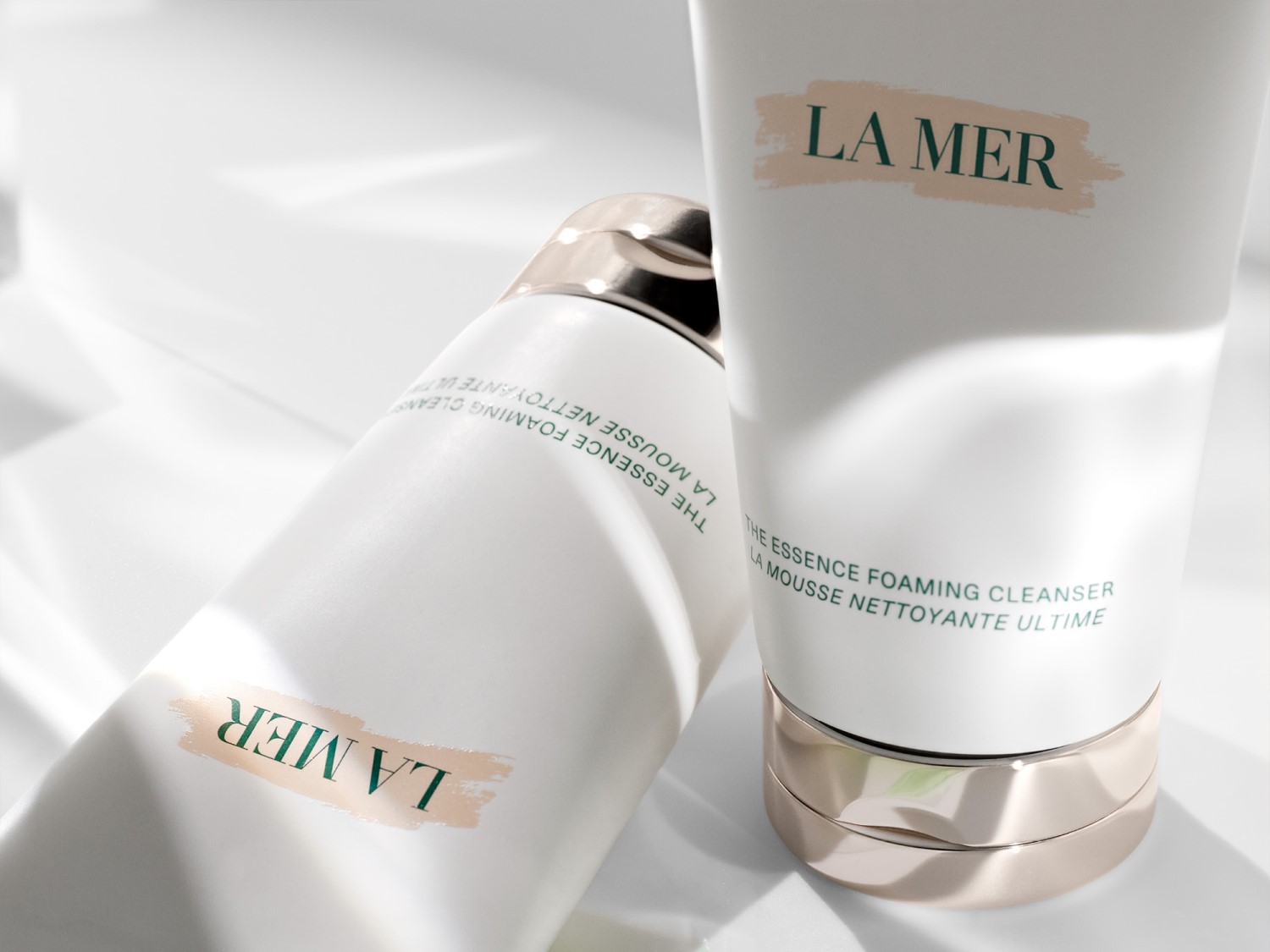 Dwa białe opakowania La Mer The Essence Foaming Cleanser z zielonymi napisami i złotymi akcentami, leżące na białym tle.