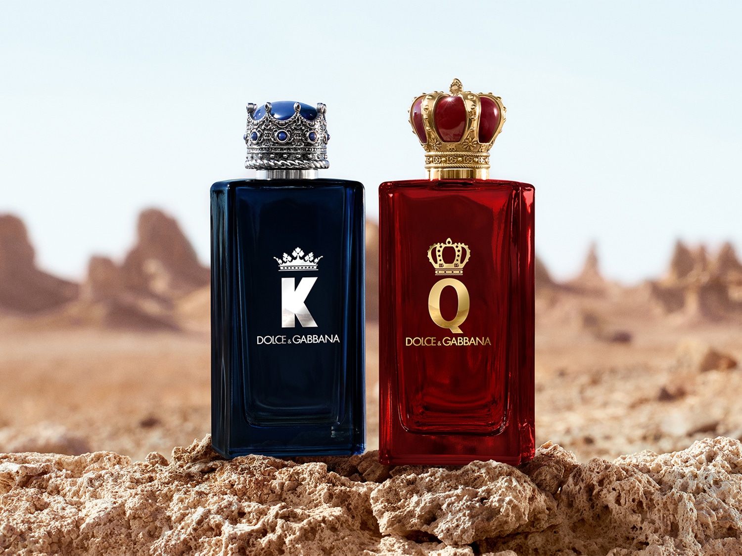 Dwa flakony perfum Dolce & Gabbana: męski, granatowy z srebrną koroną i literą K, oraz damski, czerwony ze złotą koroną i literą Q, stojące na tle pustynnego krajobrazu.
