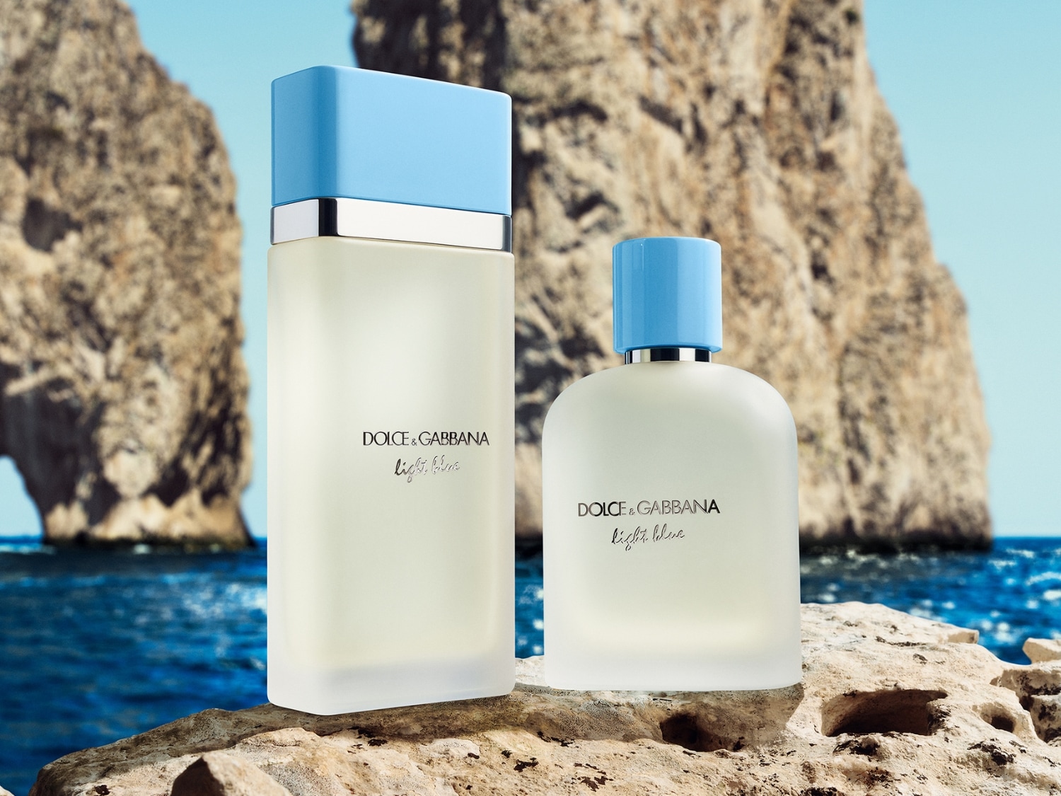 Dwa flakony perfum Dolce & Gabbana Light Blue stoją na kamieniu z widokiem na morze, większy flakon ma kształt prostokąta z jasnoniebieską nakrętką, a mniejszy jest zaokrąglony z podobną nakrętką.