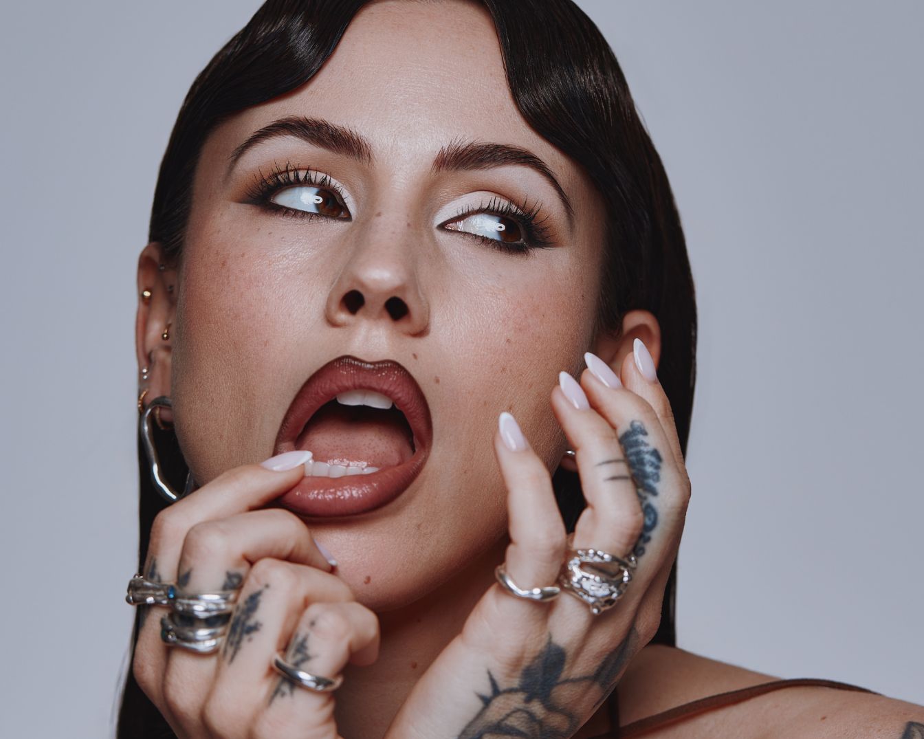 Gros plan sur une femme avec un maquillage des yeux audacieux, des lèvres brillantes et des bagues argentées originales, ajoutant une touche d'éclat à son look.