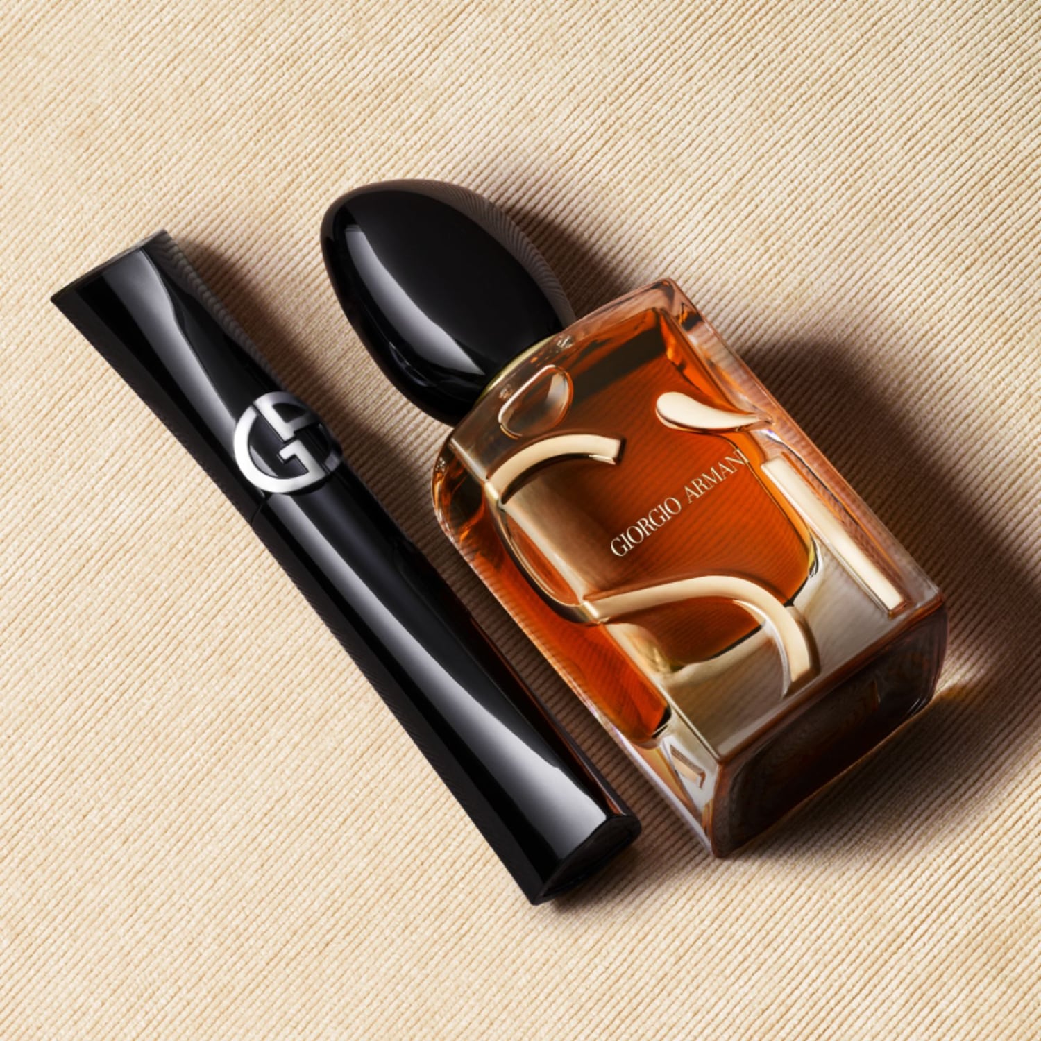 Een rechthoekige Giorgio Armani parfumfles met amberkleurige vloeistof en een zwarte mascara met het Giorgio Armani logo op een beige achtergrond.