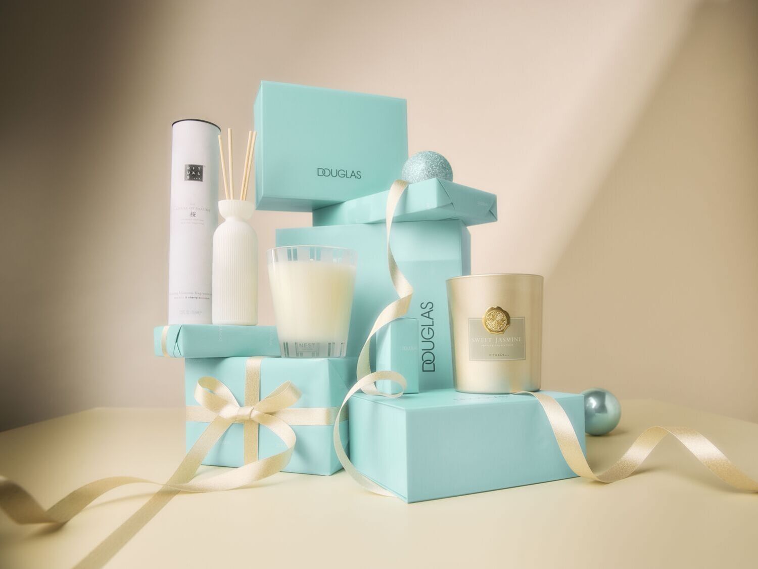 Šventinė kompozicija su „Douglas“ dovanų dėžutėmis, žvakėmis ir kvapų difuzoriumi, papuošta auksinėmis juostelėmis.