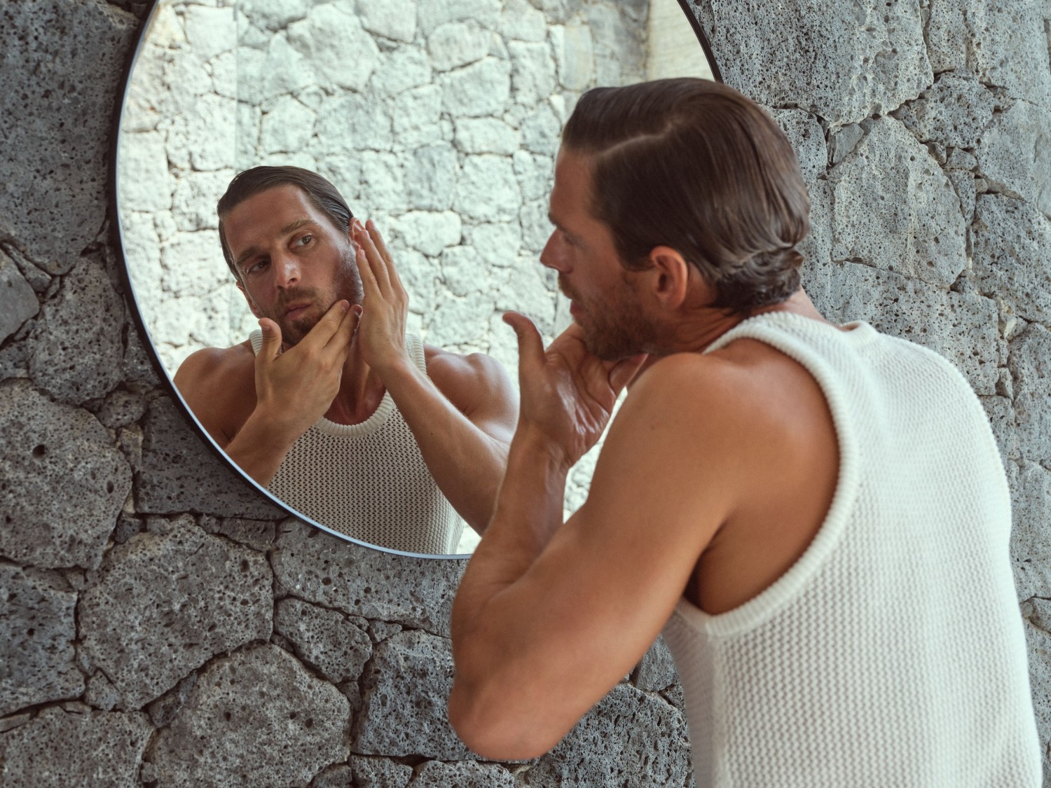 Ein Mann im weißen Tanktop betrachtet sich im Spiegel und berührt sein Gesicht, während er vor einer grauen Steinwand steht.
