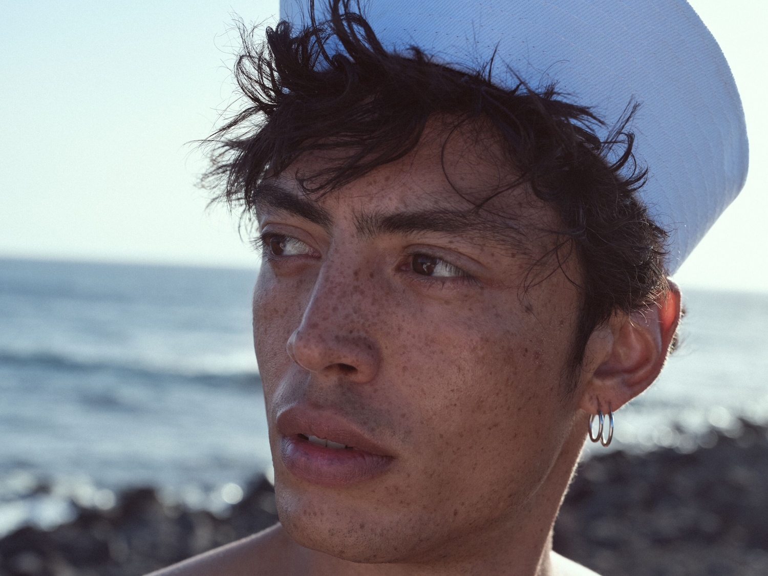 Ein junger Mann mit Sommersprossen und dunklen, lockigen Haaren trägt eine hellblaue Mütze und silberne Creolen vor einem verschwommenen Hintergrund aus Meer und Himmel.