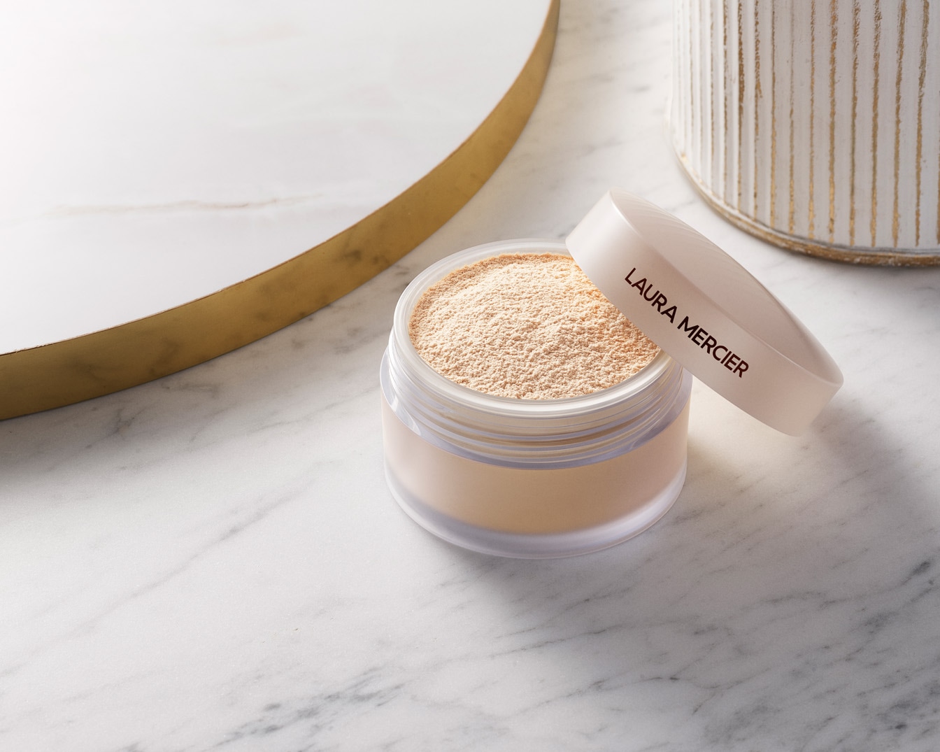 A Laura Mercier átlátszó púderének nyitott tégelye látható, finom, laza púderrel, amely tökéletes a smink rögzítéséhez és a bőr mattításához.