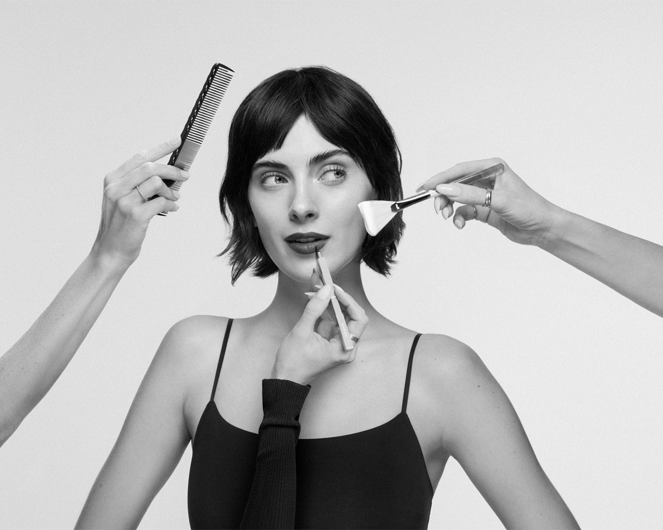 In un'immagine in bianco e nero, una donna con i capelli scuri e la canottiera nera viene truccata da diverse mani che le applicano rossetto, fard e le sistemano i capelli.