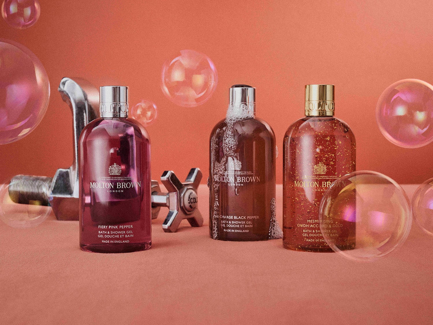 Három Molton Brown tusfürdő, köztük a Fiery Pink Pepper, egy csillogó csaptelep és lebegő buborékok előtt, rózsaszín háttérrel.