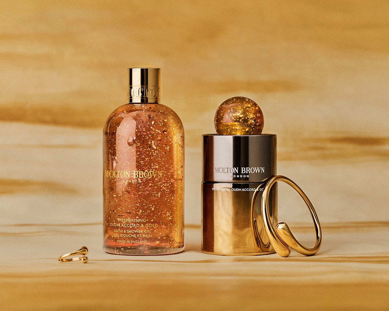 A Molton Brown fürdő- és tusfürdő gél arany színben csillogó részecskékkel, mellette pedig egy Molton Brown kölni látható, melyek az "Oudh Accord & Gold" kollekció részei.
