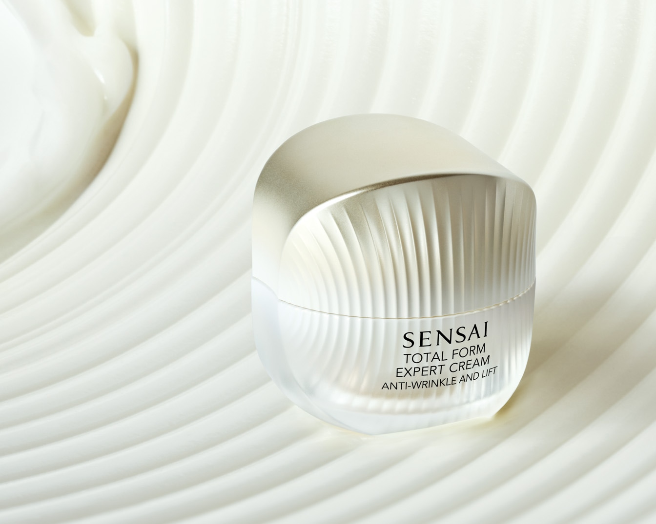 A Sensai Total Form Expert Cream egy elegáns, ezüstös kupakkal és átlátszó tégellyel rendelkezik, amely egy fehér, hullámos felületen helyezkedik el.