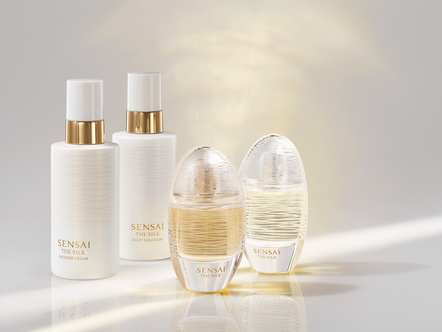 A képen a Sensai The Silk termékcsaládjának termékei láthatók: tusfürdő, testemulzió és parfüm, elegáns, fehér és arany színű csomagolásban.