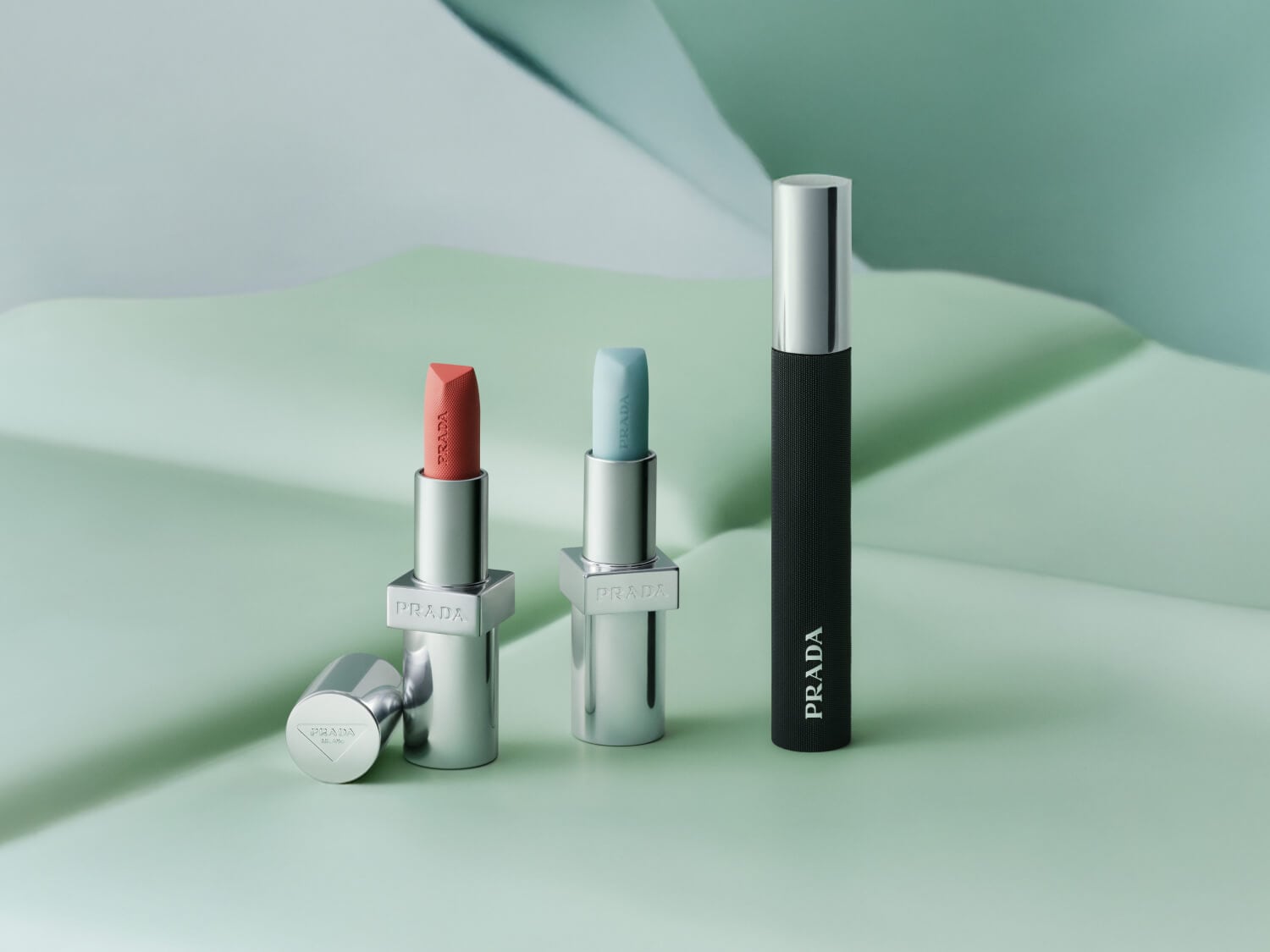 Entdecke Prada Beauty: Zwei Lippenstifte in Silberhüllen mit korallfarbenem und hellblauem Farbton, sowie eine schwarze Mascara mit silbernem Deckel.