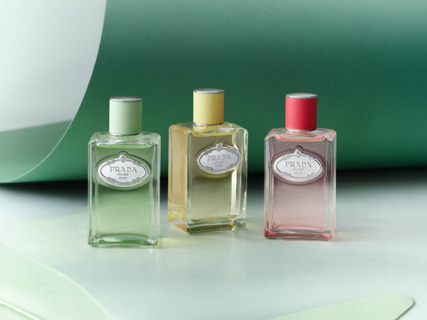 Drei Prada-Parfümflaschen in Grün, Gelb und Rosa stehen vor einem grün-weißen Hintergrund.