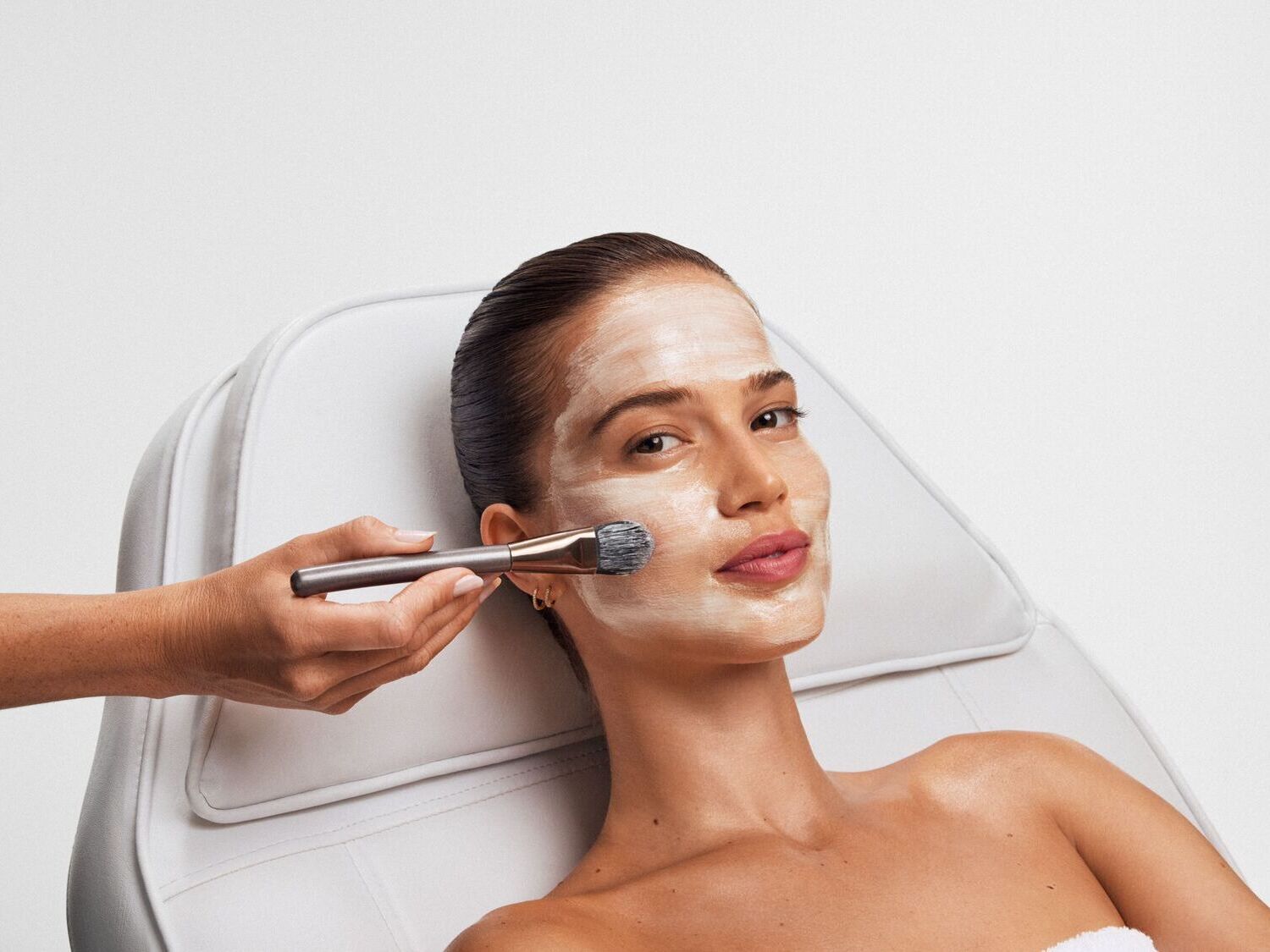 Švelnus baltas veido kremas tepamas šepetėliu ant moters veido, ji atsipalaidavusi guli ant baltos odinės kėdės.