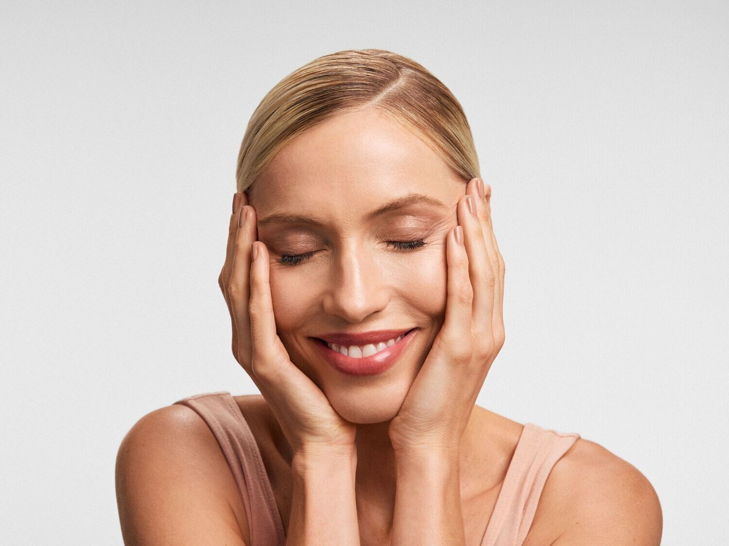 Šviesiaplaukė moteris užmerktomis akimis šypsosi, rankomis prilaikydama veidą, spinduliuoja ramybe ir pasitenkinimu.