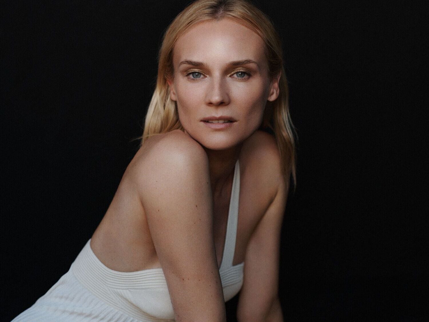 Šviesiaplaukė moteris su auksiniu akių makiažu ir balta suknele pozuoja priešais juodą foną.