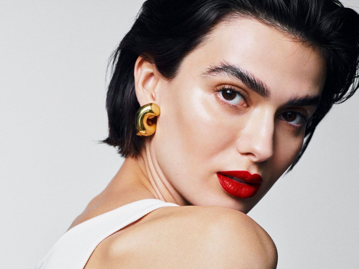 Moters portretas su ryškiai raudonu lūpdažiu, masyviais auksiniais auskarais ir trumpais juodais plaukais, žvelgiančios per petį.