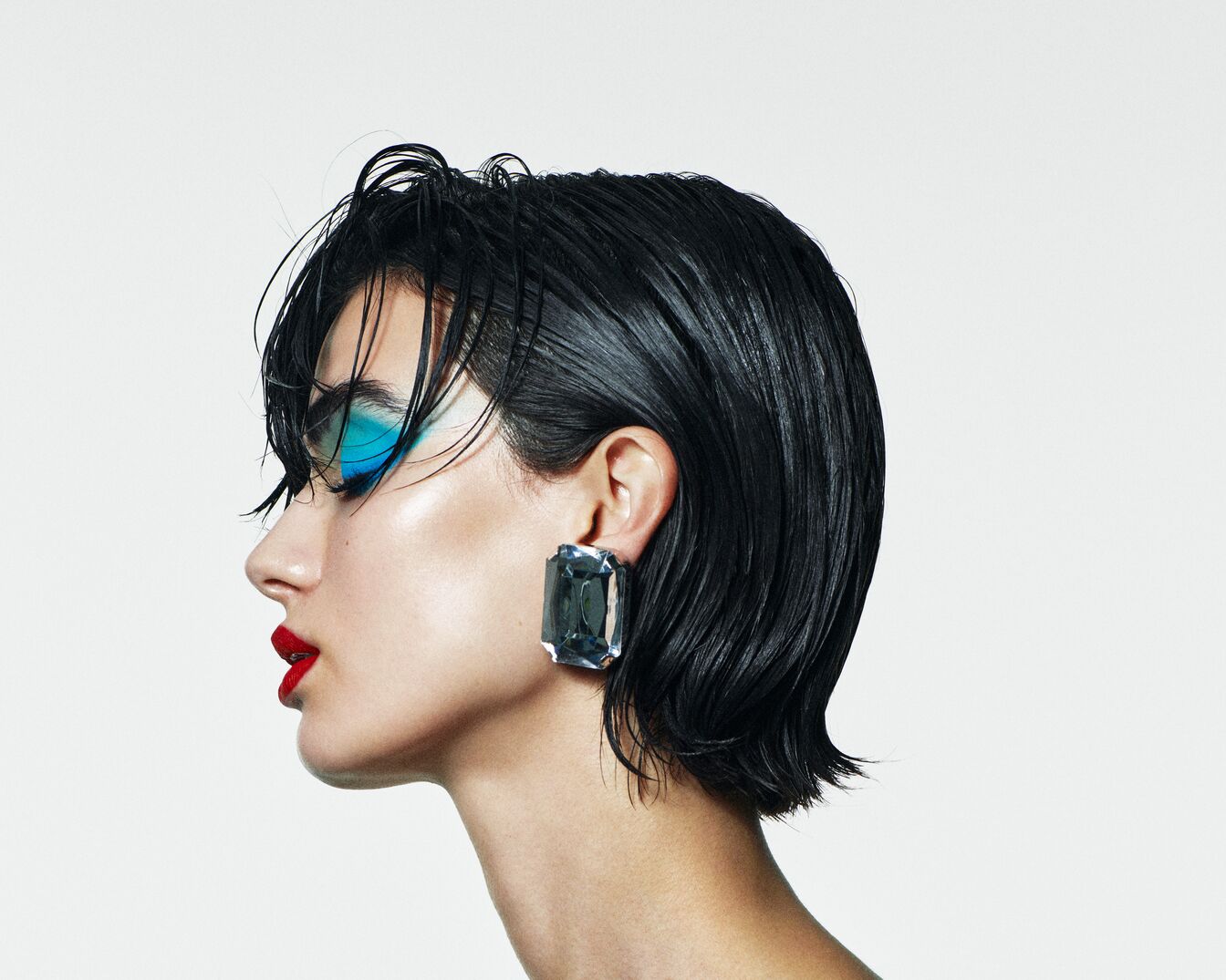 Šoninis moters portretas su trumpais juodais plaukais, ryškiai mėlynais akių šešėliais, raudonais lūpų dažais ir dideliu stačiakampiu auskaru.