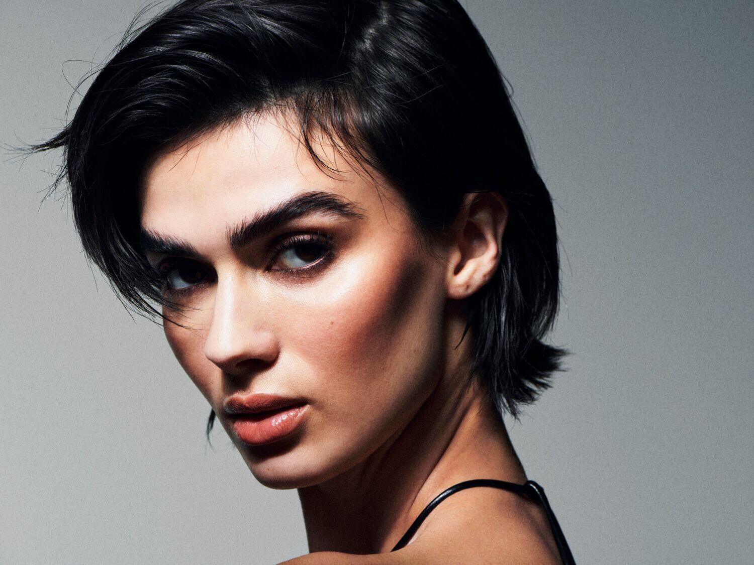 Portretas moters su trumpais juodais plaukais, ryškiu makiažu ir blizgančiomis lūpomis, žiūrinčios į kamerą.