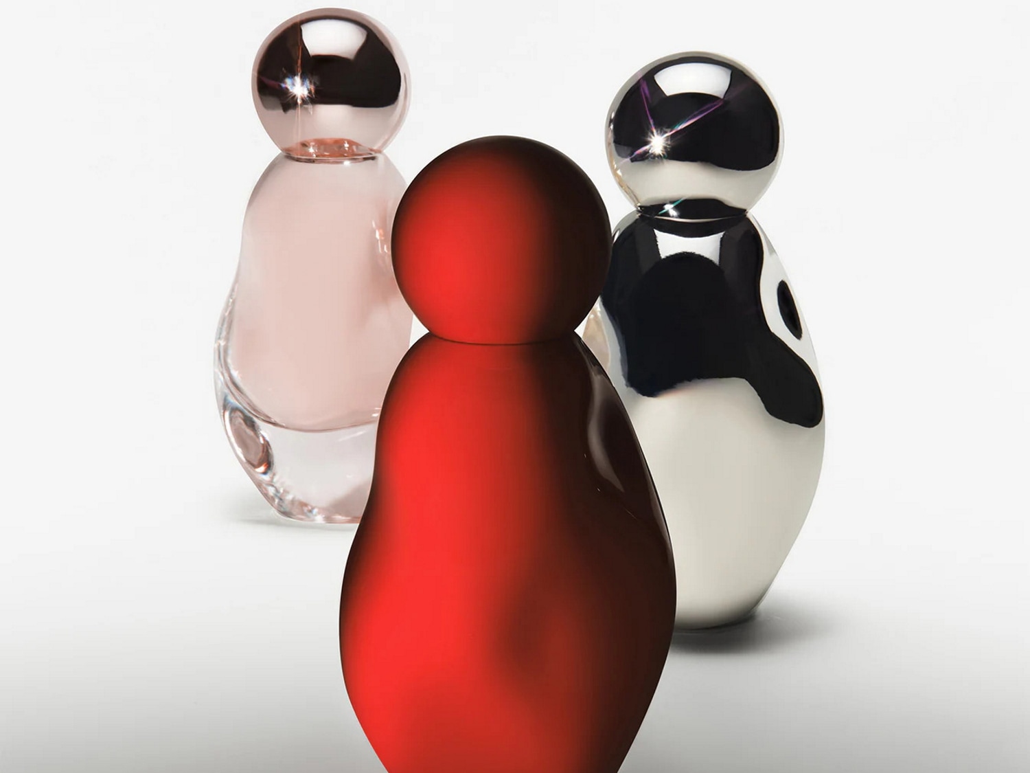 Trois flacons de parfum aux formes arrondies et aux couleurs vives : rouge, rose pâle et argenté, chacun coiffé d'un bouchon sphérique brillant.