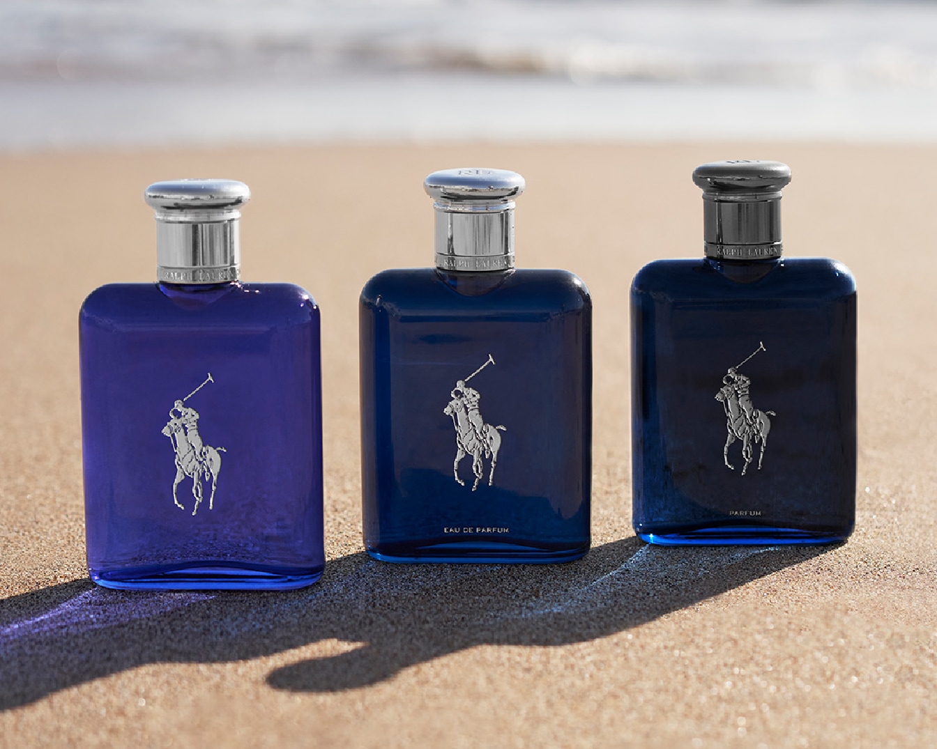 Trois flacons de parfum Ralph Lauren bleu marine, violet et bleu foncé sur une plage de sable, chacun orné du logo emblématique du joueur de polo.