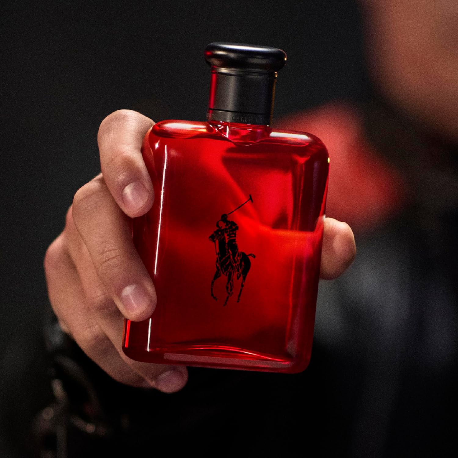 Gros plan sur un flacon de parfum rouge vif Ralph Lauren, orné du logo emblématique du joueur de polo noir, tenu délicatement dans une main.