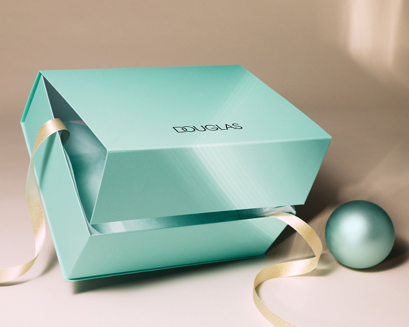 Geöffnete, helltürkise Douglas-Geschenkbox mit goldener Schleife und passender Kugel, die Eleganz und Vorfreude auf den Inhalt verspricht.