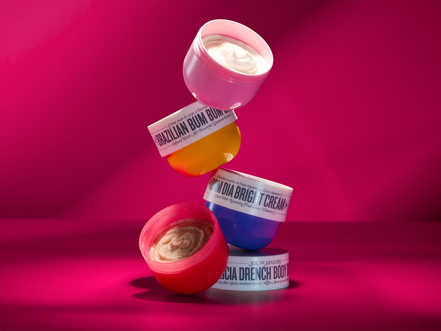 Spalvingas „Sol de Janeiro“ kūno kremų rinkinys, įskaitant „Brazilian Bum Bum Cream“, „Bom Dia Bright Cream“ ir „Cica Drench Body Cream“, sukrautas ant ryškiai rožinio fono.