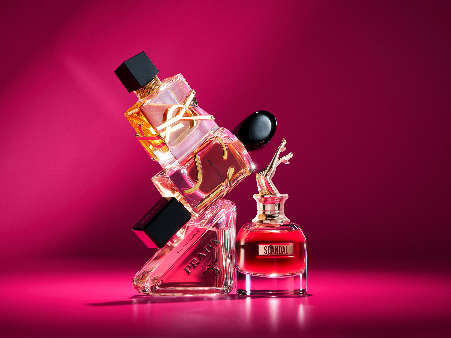 Négy különböző parfümös üveg egymásra helyezve, köztük YSL, Giorgio Armani, Prada és Scandal, mindegyik rózsaszín háttér előtt.
